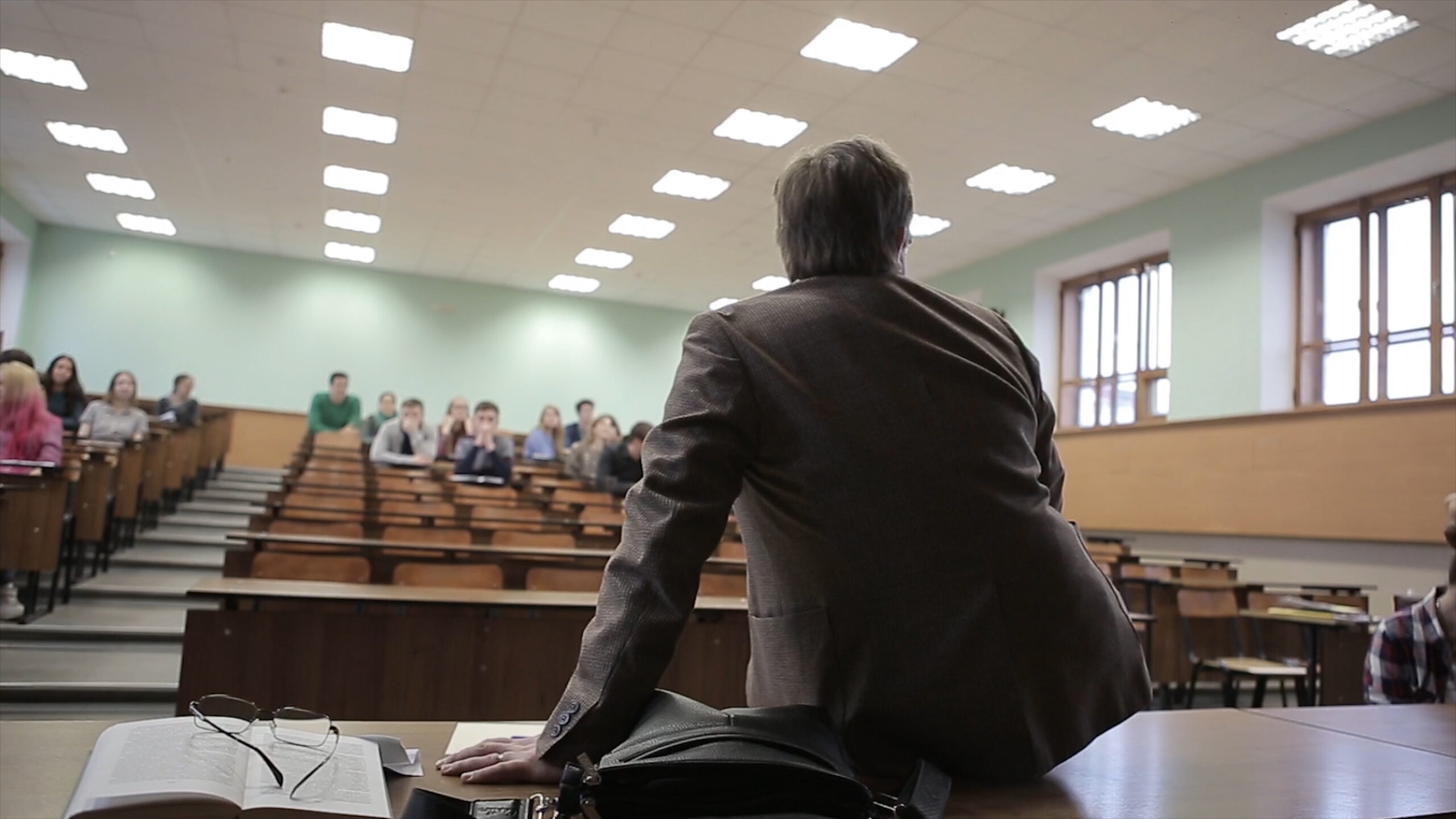 Back view of man presenting to students at a lecture theatre. View from the back of the teacher who is sitting on the table.
